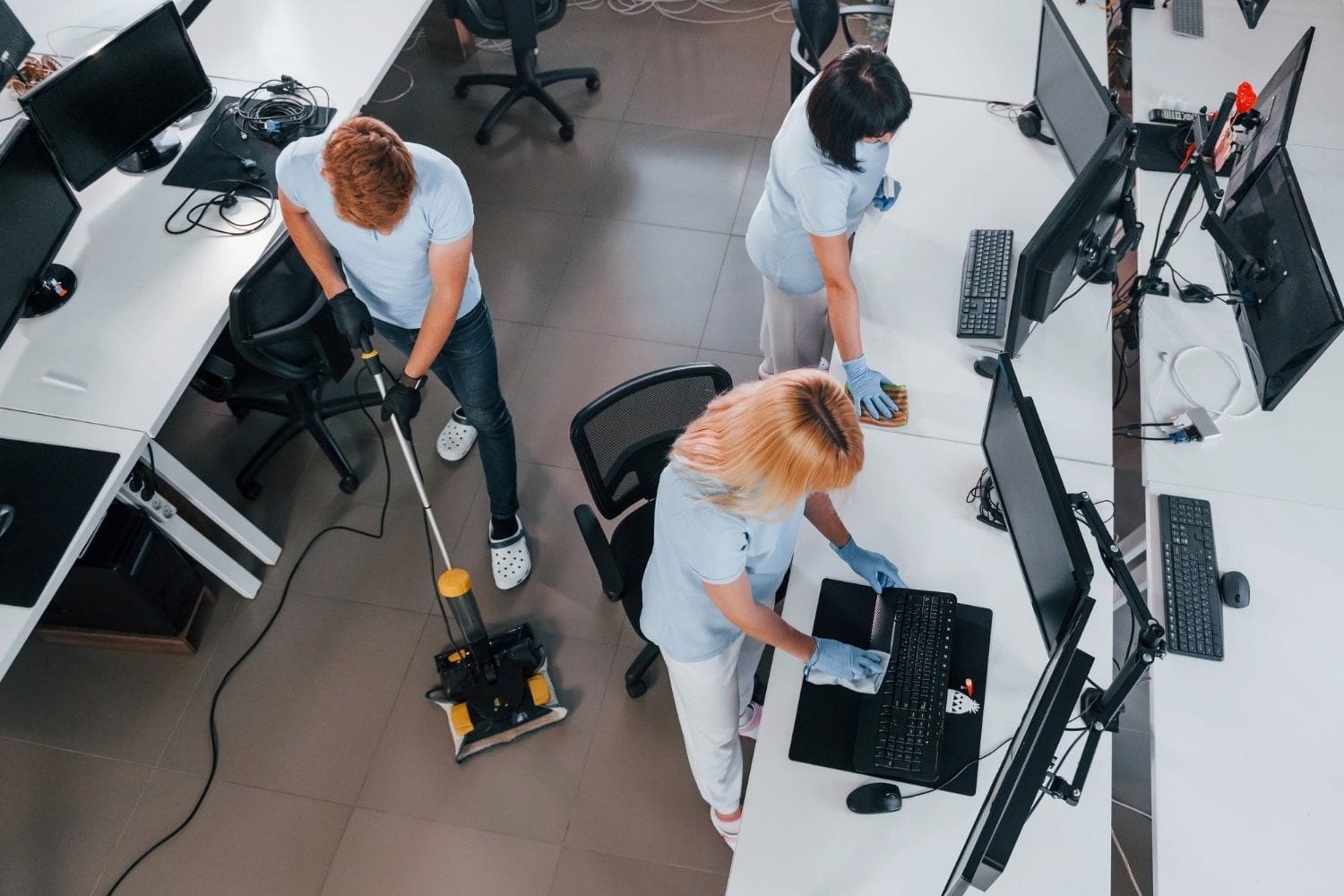 People working with computers at desks in an office, one vacuuming the floor