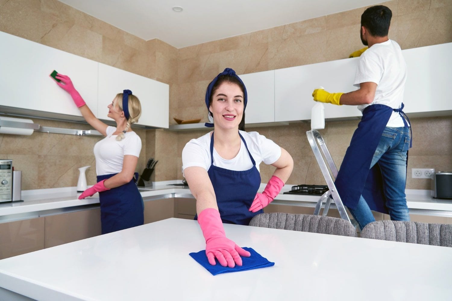 Three cleaners in pink gloves cleaning a bright kitchen with sponges and spray bottles.