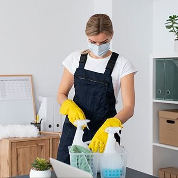 Person in yellow gloves and mask cleaning a desk with spray bottles in an office setting