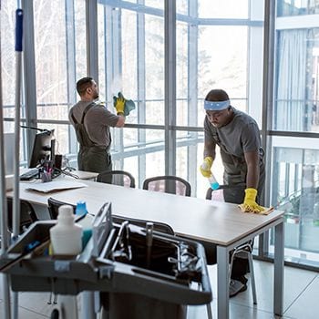 Two workers in gloves cleaning a modern office conference room near large windows.