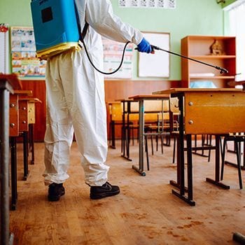 Person in protective suit spraying disinfectant in a classroom with desks and chairs.